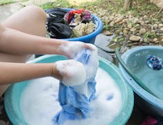 A person crouched over a soapy bucket of water hand washing their clothing.