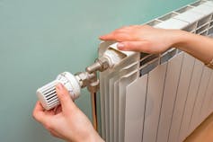 A person's hands adjusting a thermostatic radiator valve against a blue wall.