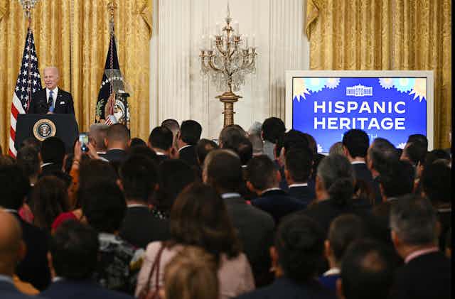 Biden at a lectern in front of a crowd, with a screen reading 'Hispanic Heritage'