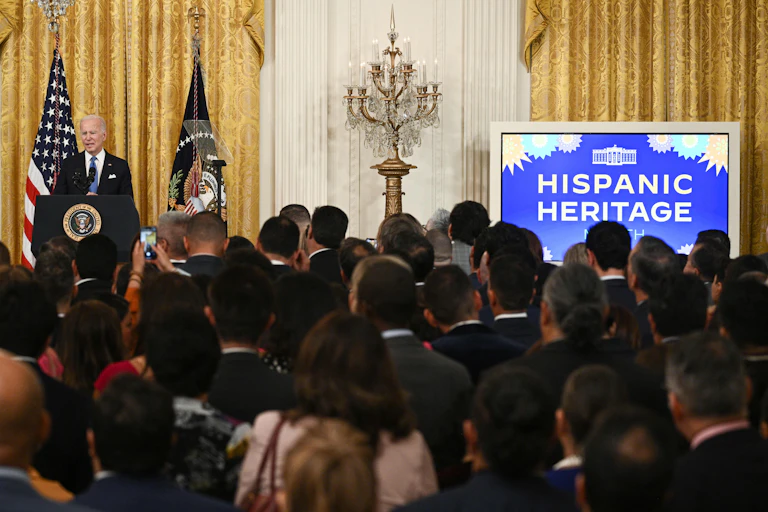 Biden at a lectern in front of a crowd, with a screen reading 'Hispanic Heritage'