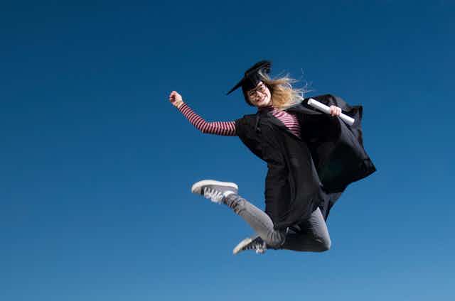 A young woman graduate in hat and gown jumping for joy against a blue sky.