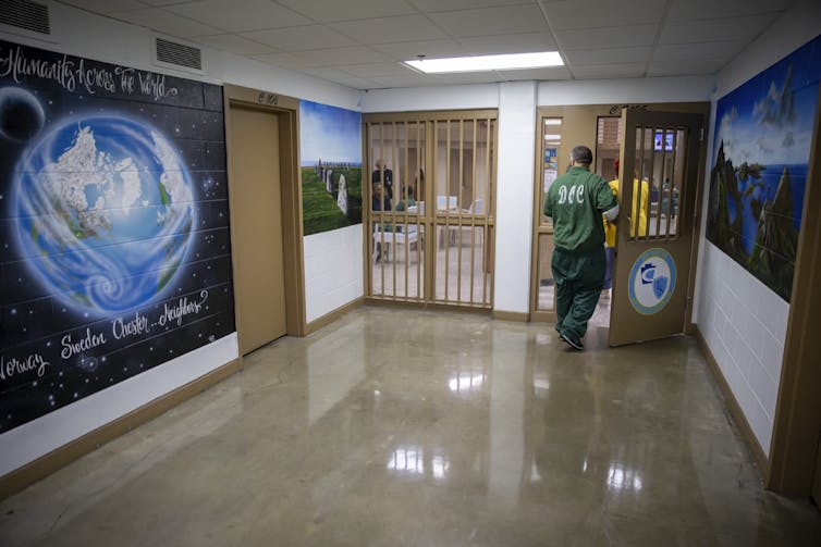 A man in a green outfit walks past a large mural of a world in an indoor hallway.