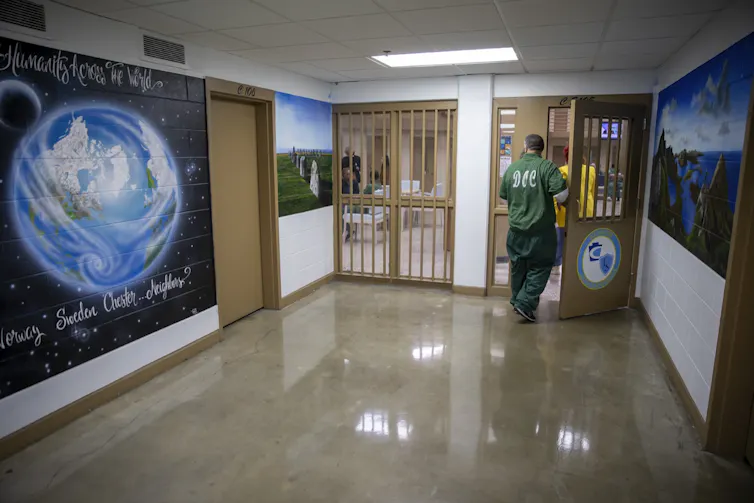 A man in a green outfit walks past a large mural of a world in an indoor hallway.