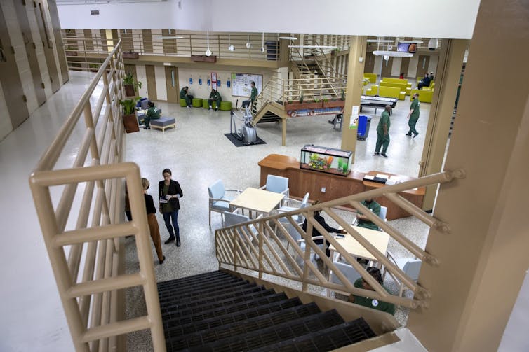 An overhead shot shows an open looking common room with some people in green prison uniforms sitting and standing.