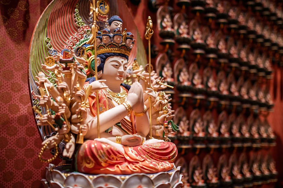 Statue of a richly dressed seated deity with many arms, two of which are joined in prayer.