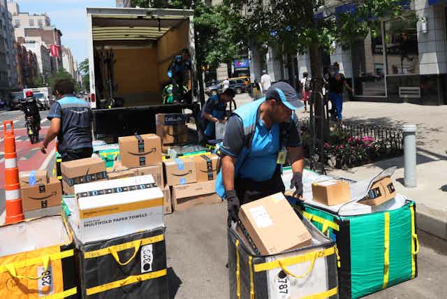 Delivery workers surrounded by dozens of packages piled behind an open truck on a city street