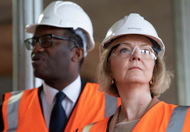 A white woman and a black man, both wearing white hard helmets, eye coverings and orange safety vests, look in opposite directions 