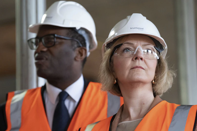 A white woman and a black man, both wearing white hard helmets, eye coverings and orange safety vests, look in opposite directions