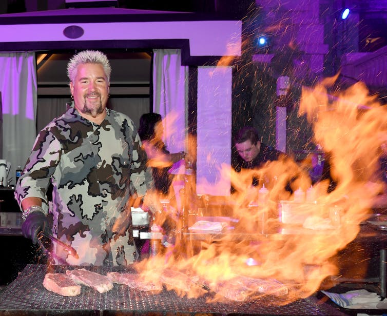 A bleached blond man with a goatee and wearing a camo shirt grills steaks over a fire, which is flaming high.