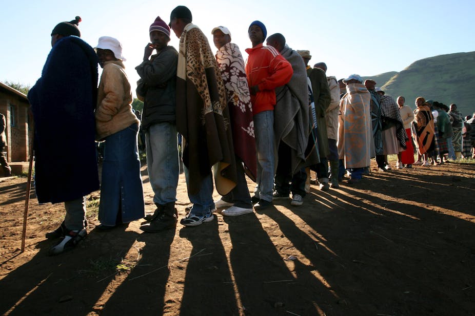 Men and women, some wearing traditional Basotho blankets, stand in line to vote.