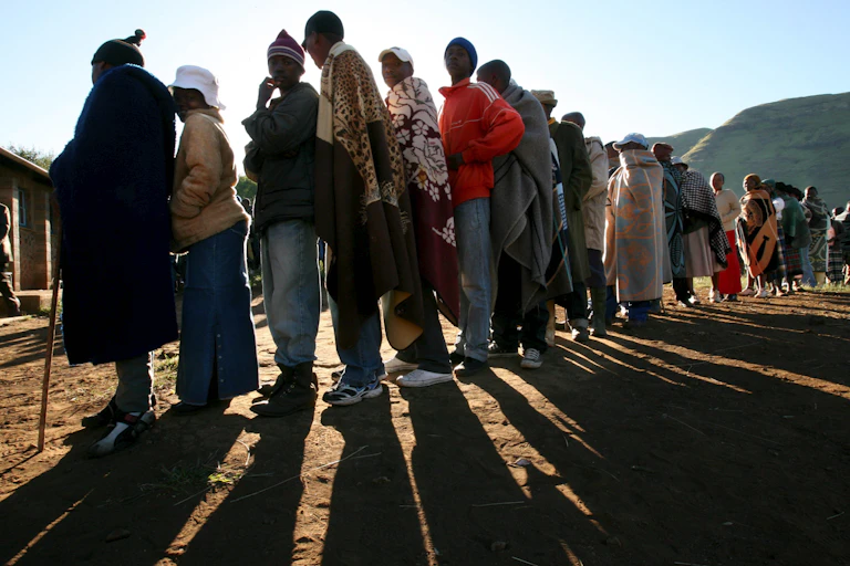 Men and women, some wearing traditional Basotho blankets, stand in line to vote.