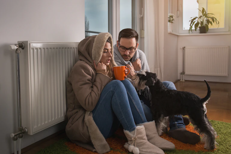 A man a woman sat on the floor next to a radiator with their dog trying to stay warm