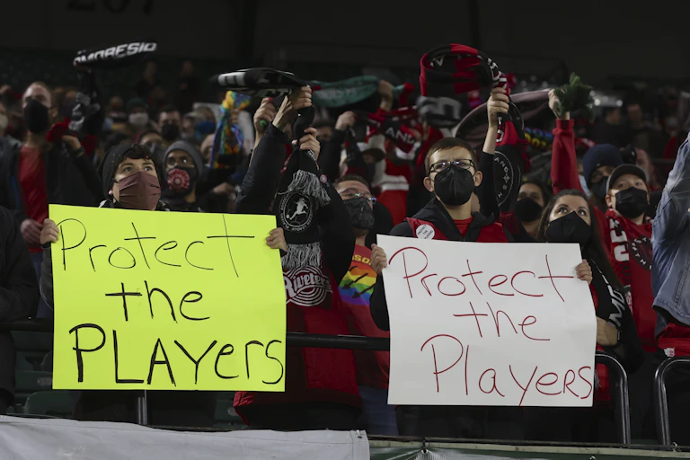 A crowd of football supporters holding scarfs, with two at the front holding placards stating 'Protect the Players'