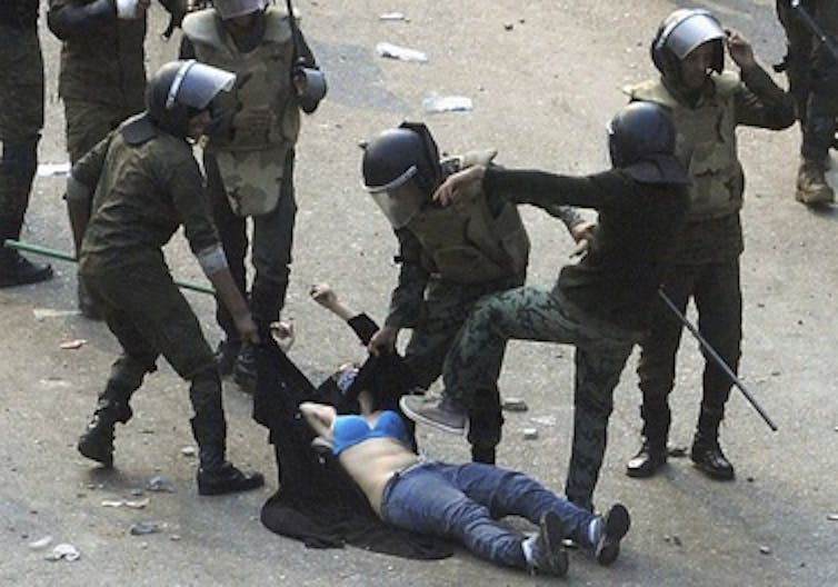 A half clothed woman wearing a blue bra and jeans being dragged by policemen, with one about to step on her.