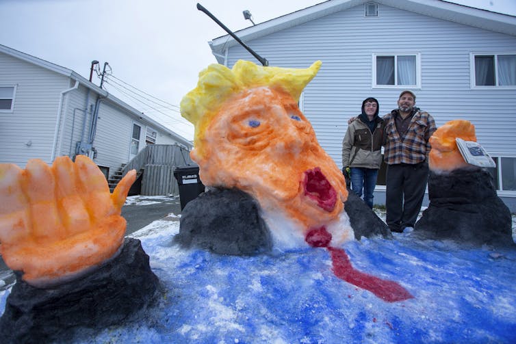 A teenaged boy and a man pose for a photo with a snow sculpture that depicts a yellow-haired man wearing a red tie melting into the water.