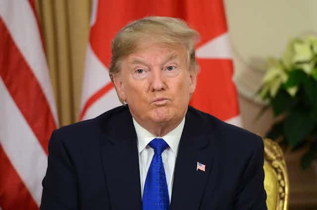 A blond man in a blue suit and tie is shown sitting in a gilded chair with the Canadian flag behind him.