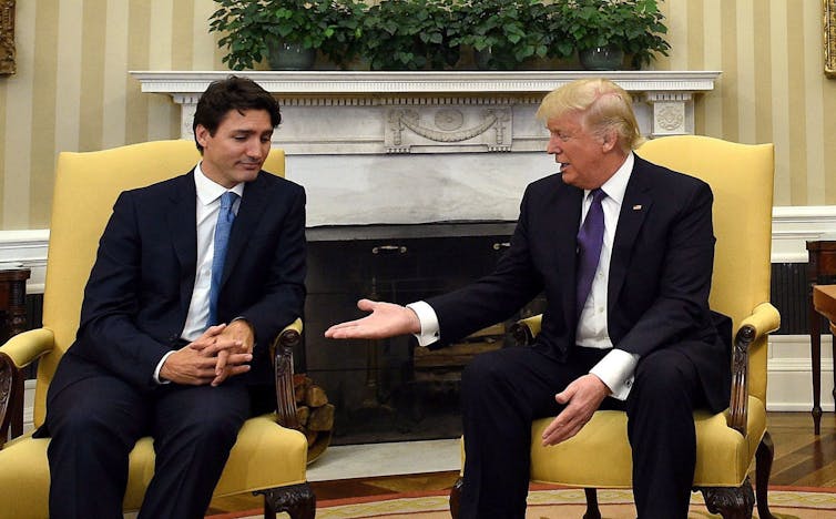 A dark-haired man looks down skeptically at the outreached hand of an older blond man sitting next to him in an ornate office.