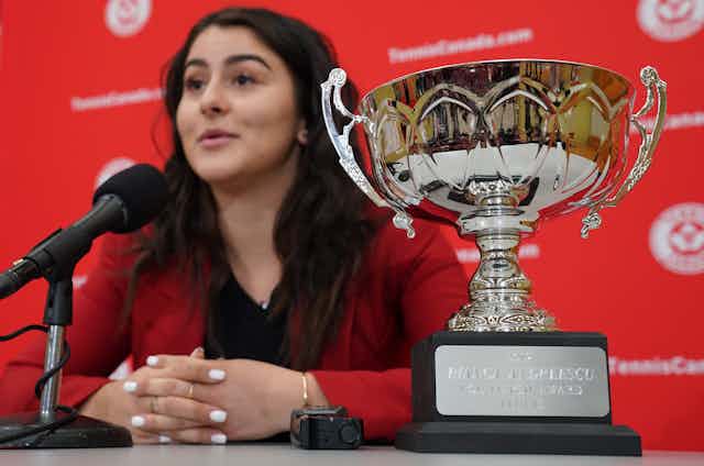 A large trophy cup sits on a table beside a dark-haired woman in a red blazer 