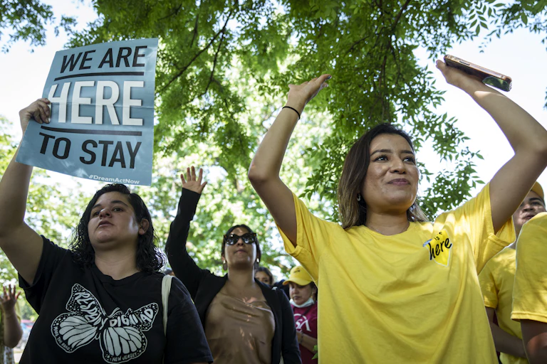 Three young, Latina women hold up their hands and one holds a sign that says 'We are here to stay.'