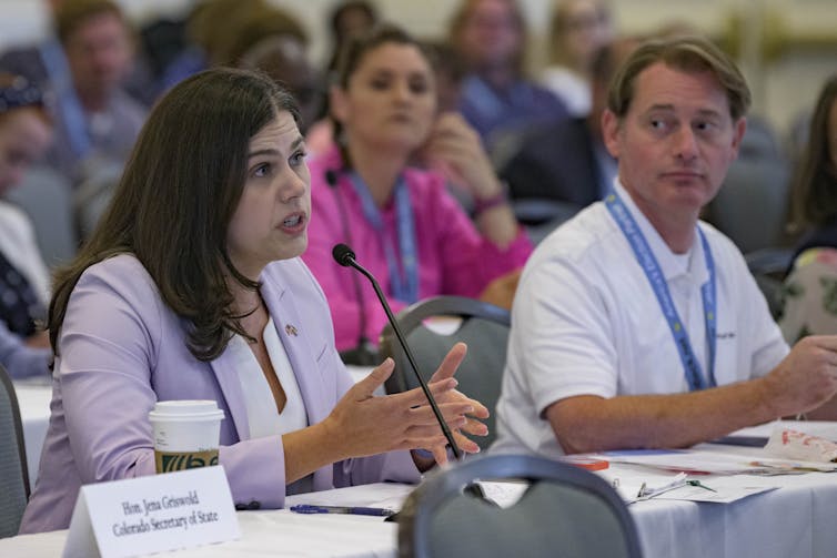 A woman with dark hair and wearing a lavender jacket talks into a microphone at a meeting with many people.