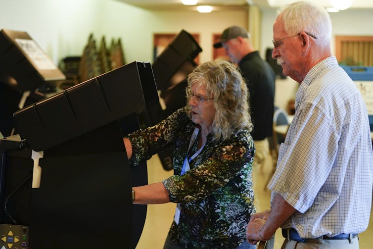 A woman with gray hair helps a man with gray hair cast a ballot at a voting machine.