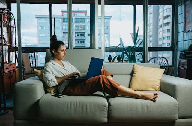 Young woman in an apartment reclines on a couch, scrolling on her laptop