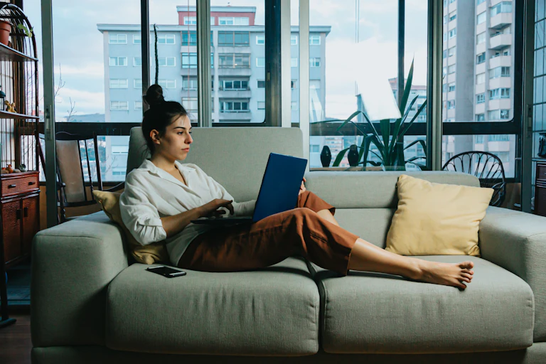 Young woman in an apartment reclines on a couch, scrolling on her laptop