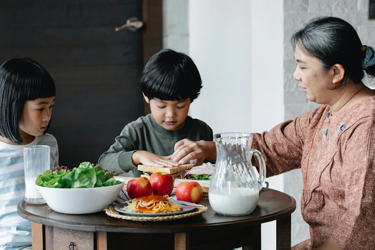Two siblings and an older woman sit down to a table of food.