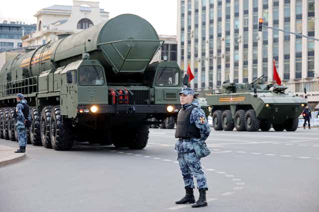 A Russian soldier stands in the street as a strategic nuclear warhead passes in the background.