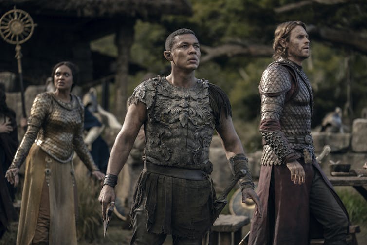 An elf looking watchful and alert with pointed ears in body armour stares into the distance flanked by two other figures.