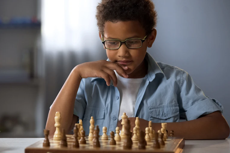 A young boy sits at a table focusing on a wooden chess board with pieces.