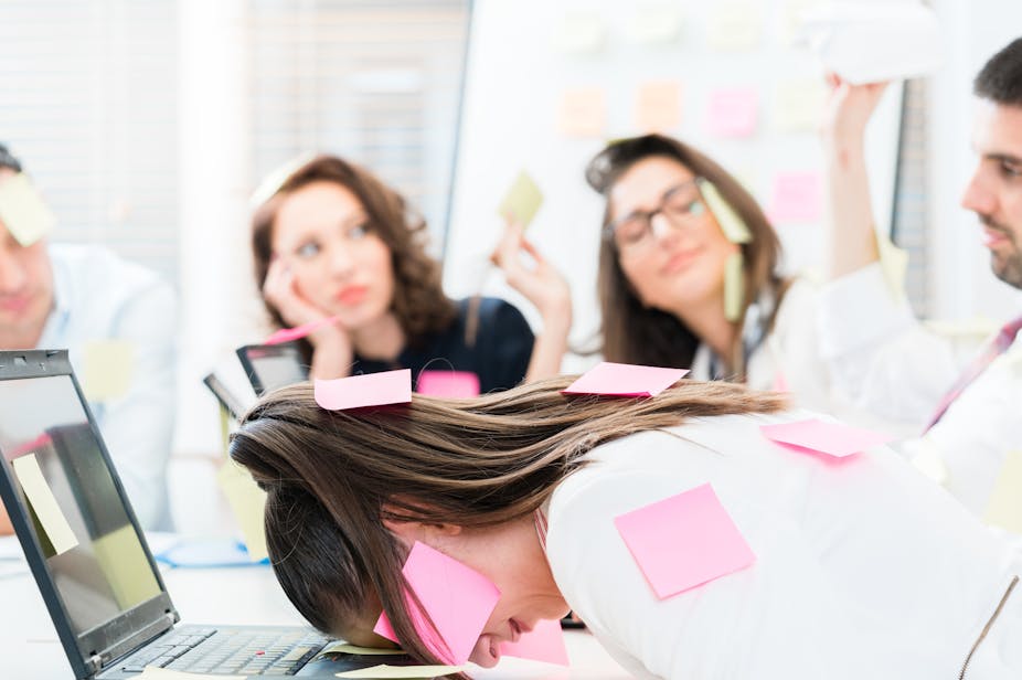 Woman covered in post-its banging head on laptop surrounded by colleagues.