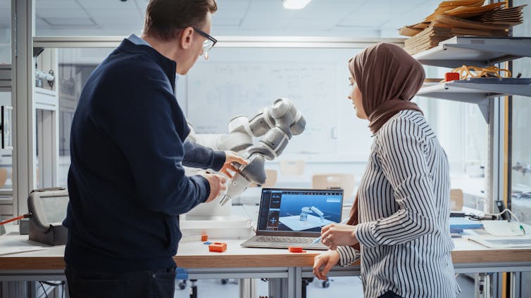 A man holding a robotic arm and talking to a woman in a hijab in front of a laptop screen