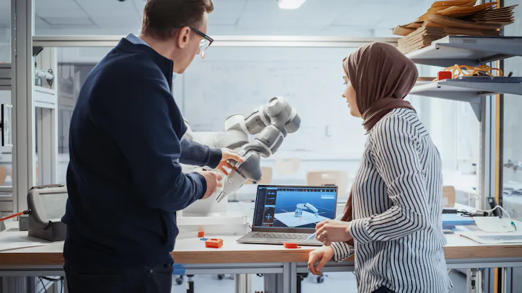 A man holding a robotic arm and talking to a woman in a hijab in front of a laptop screen