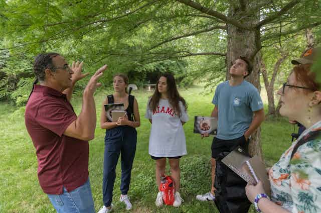A man holds up his hands while speaking to a group of college students standing on grass under a tree.