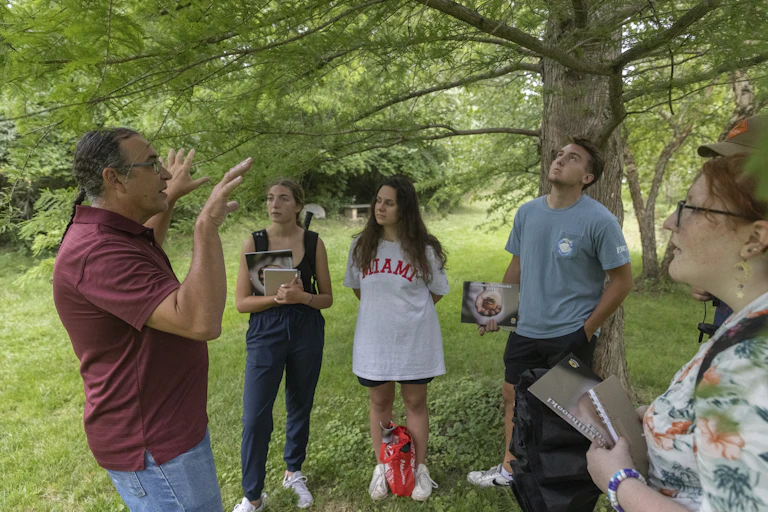 A man holds up his hands while speaking to a group of college students standing on grass under a tree.