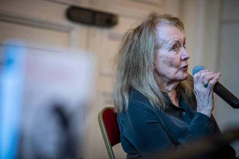 A beautifully groomed 84-year-old woman speaks seriously in a microphone