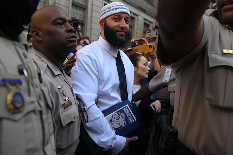 A man in a white shirt and taqiyah surrounded by police officers in khaki uniforms.