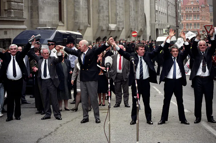 Six men in black suits hold up hands in triumph in the street.