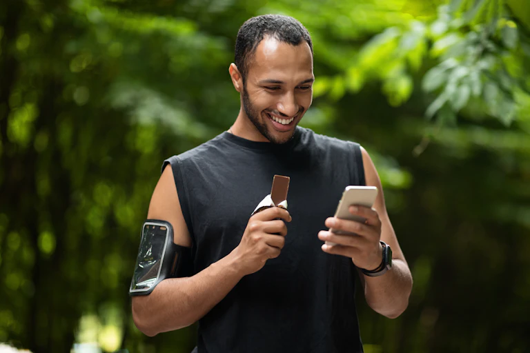 A man takes a break from his workout outside to look at his phone and eat a chocolate bar.