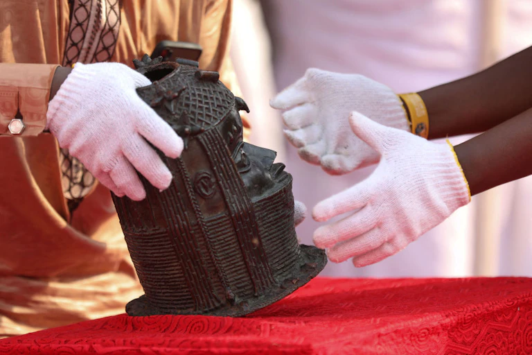 a sculpture of an adorned human head is placed on a red cloth by hands in white gloves.
