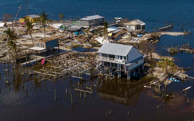A house on stilts stands amid wrecked homes with water surrounding them.