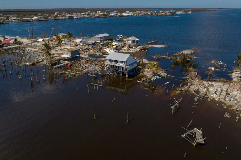 A house on stilts stands amid wrecked homes with water surrounding them.
