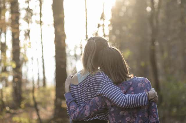 A view from behind of two women with their arms around each other outdoors. 