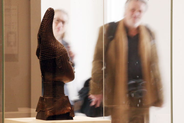 A statue of a head behind a glass display case, in the background a man and woman gaze at it.