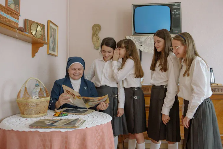 Unas niñas de uniforme observan a una monja leer una revista.