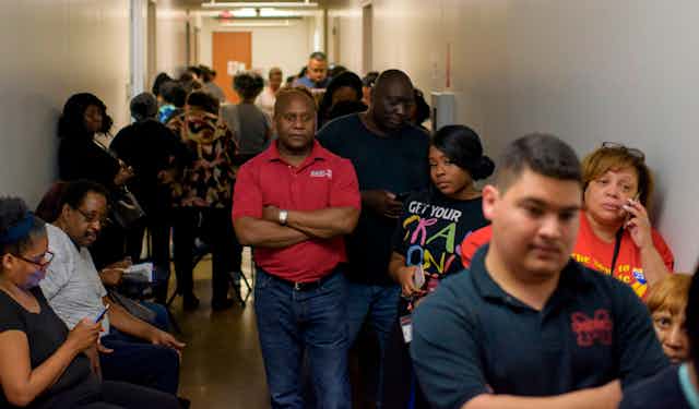 A black man in a red shirt has his arms crossed as he waits with hundreds of other voters to cast their ballot during the presidential primary.