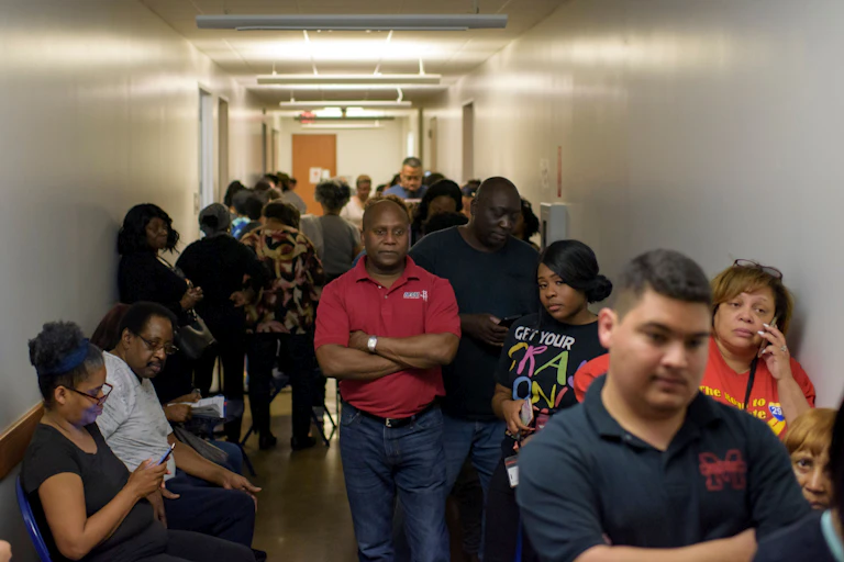 A black man in a red shirt has his arms crossed as he waits with hundreds of other voters to cast their ballot during the presidential primary.