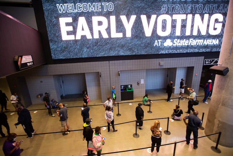 A gigantic banner with the words Early Voting hangs on a wall above dozens of people waiting to vote.