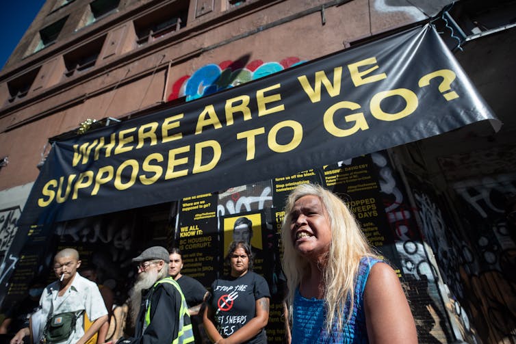 A group of people stand in front of a brown building carrying a banner that reads: where are we supposed to go?